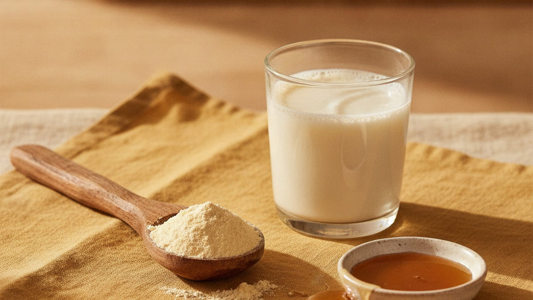 A wooden spoon of pale colostrum powder beside a glass of fresh milk and a dish of honey on warm linen